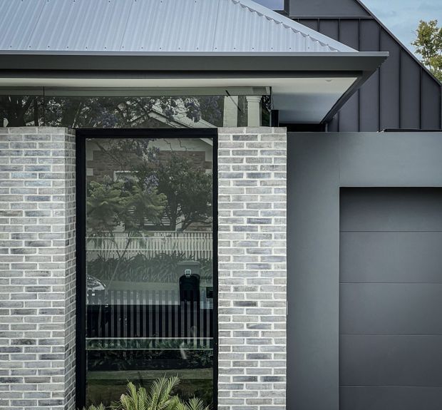 Close-up exterior view of a contemporary residential facade featuring pale grey brickwork, a large black-framed glazed entry door, dark grey metal cladding and garage door, and a light grey corrugated metal roof with a broad projecting eave. Reflections reveal trees and a front fence beyond the glazing.