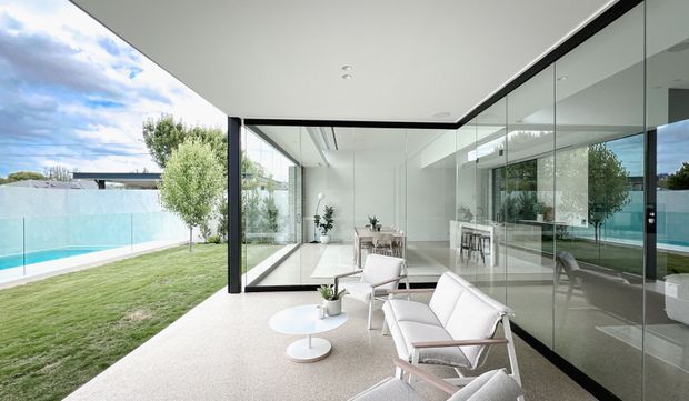A bright contemporary indoor-outdoor living area with floor-to-ceiling glazing opening onto a lawn and swimming pool. The minimalist white interior contains a dining table and chairs, while the foreground has a small lounge setting with white upholstered armchairs and round tables. Slim dark window frames and extensive glass create strong visual continuity between the interior and landscaped exterior.