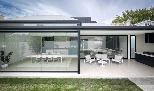 Contemporary single-storey home extension with extensive floor-to-ceiling glazing opening onto a landscaped lawn. The enclosed dining area and adjacent outdoor lounge are arranged beneath a broad low-pitched roof, with pale terrazzo-like flooring, dark slim window frames, and a compact outdoor kitchen or barbecue zone at right. A swimming pool and outdoor furniture are visible through the glazing.