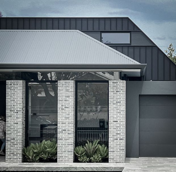 Front exterior view of a contemporary single-storey residence with a dark charcoal metal-clad upper gable, light grey corrugated metal roof, pale brick facade, black-framed floor-to-ceiling glazing, and an integrated dark garage door. Low architectural planting lines the entry glazing and a paved driveway sits in the foreground.