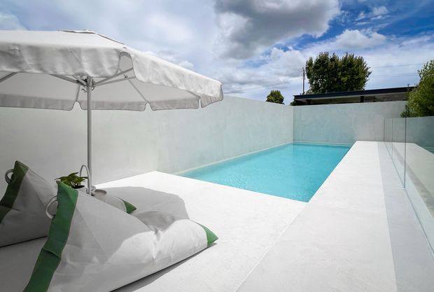 A minimalist outdoor pool courtyard with a bright turquoise rectangular swimming pool, white rendered boundary walls and pale paving. A large white sun umbrella shades oversized white and green beanbag loungers in the foreground, while a frameless glass balustrade lines the right side. The scene is bright and contemporary with strong white surfaces and a partly cloudy sky.