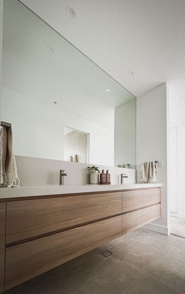 Bright, minimalist bathroom featuring a long floating timber vanity with integrated double basins, brushed metal tapware, a pale stone or solid-surface benchtop and matching half-height wall finish. A large wall-to-wall mirror spans above the vanity, with warm neutral towels, simple bottles and a small plant adding restrained styling. The floor is finished in large grey-beige tiles, and recessed ceiling lights provide even illumination.