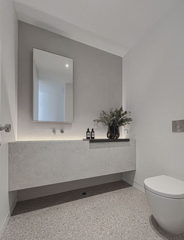 A minimalist contemporary bathroom featuring a wall-mounted concrete-look vanity with integrated basin, frameless rectangular mirror, wall-mounted tapware, recessed shelf, and a white toilet. The room has pale grey walls, a dark under-vanity recess, terrazzo-style flooring, and understated brushed-metal hardware. A small vase and toiletries sit on the vanity shelf.