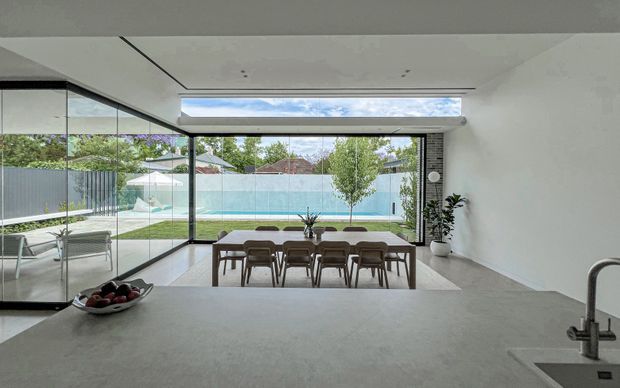 A bright, minimalist open-plan dining and kitchen area with floor-to-ceiling black-framed glazing overlooking a swimming pool and landscaped backyard. A long timber dining table with multiple chairs sits centrally, while a pale stone kitchen benchtop and undermount sink occupy the foreground. The room features a smooth white ceiling with recessed linear detailing and a long clerestory-style skylight or high window, creating an airy connection to the outdoors.