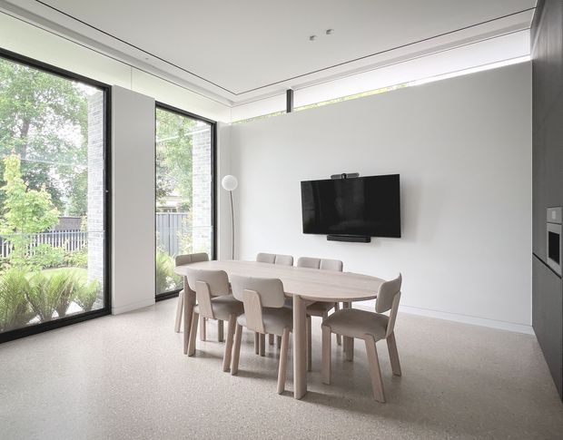 Bright, minimalist dining area with a large light-wood dining table and matching upholstered chairs, positioned in front of a wall-mounted television. Full-height black-framed glazing opens the room visually to a leafy garden, while a concealed linear ceiling detail and pale neutral finishes create a clean contemporary interior.