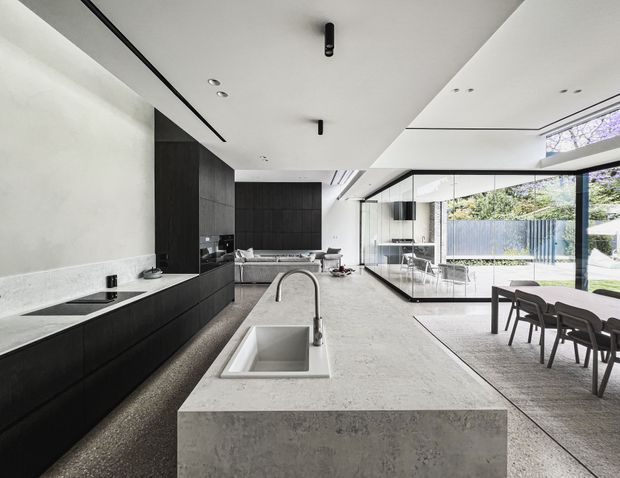 A spacious open-plan contemporary kitchen with a large pale stone island, integrated sink and curved tap, matte black full-height cabinetry, and a matching stone benchtop along the rear wall. The kitchen opens to a dining area and glazed living space, with extensive floor-to-ceiling windows connecting to a landscaped outdoor area. Recessed and minimal linear ceiling lighting, dark-framed glazing, and a restrained monochromatic palette create a refined modern interior.