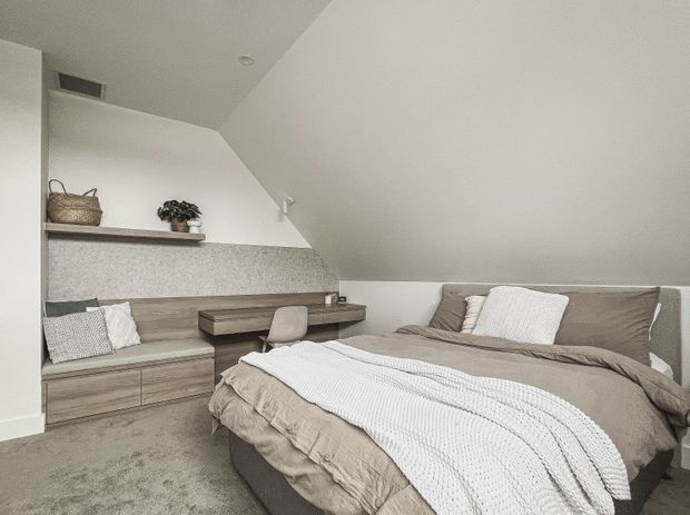 A compact attic bedroom with a sloped ceiling and dormer-like angled wall. The room features a built-in timber-toned window seat and low storage cabinetry, a matching wall-mounted desk with a small upholstered chair, open shelving with minimal decor, and a double bed dressed in taupe bedding with a white textured throw. The palette is soft neutral and the styling is restrained and contemporary.