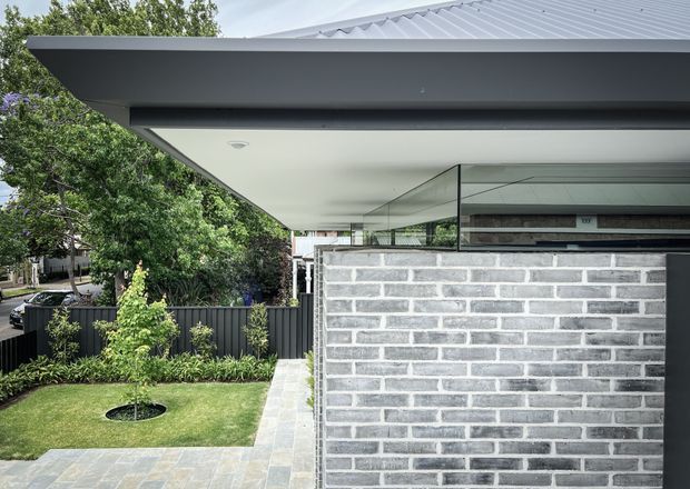 Contemporary single-storey residential exterior viewed from a landscaped side path. The image highlights a broad dark metal roof with a deep cantilevered eave, white soffit, full-height corner glazing, and a low pale grey rendered or painted brick wall. A manicured lawn, planting, and dark vertical fencing surround the building.