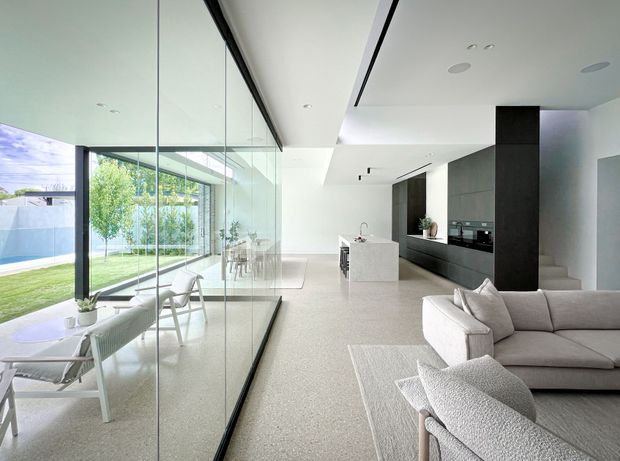 Bright open-plan contemporary interior with a living room, dining area, and kitchen arranged around extensive floor-to-ceiling glazing. A pale textured floor, white walls and ceiling, minimalist white island, and dark full-height kitchen cabinetry create a restrained modern palette. The glazed dining zone opens visually to a landscaped lawn and pool area, while a large neutral sectional sofa anchors the foreground.