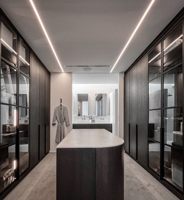 A contemporary dressing room and bathroom suite with a central island, full-height dark timber cabinetry, black-framed glass-fronted wardrobes, and integrated linear ceiling lighting. A vanity bathroom with a large mirror is visible at the rear, while a robe hangs beside the entrance. The palette is sophisticated and monochromatic, combining dark wood, soft grey surfaces, white walls, and black metal framing.