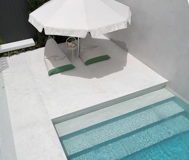 A minimalist outdoor pool area with a pale paved terrace, turquoise swimming pool, broad submerged entry steps, and a large white umbrella shading two low loungers with green accents. The setting is bright, clean, and resort-like, with simple white architectural surfaces.