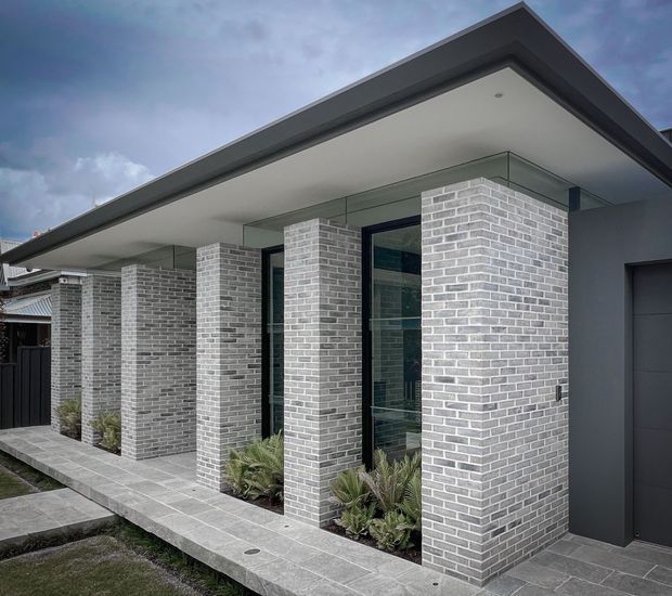 Contemporary single-storey residential facade with a broad flat roof, deep covered entry canopy, repeated pale grey brick columns, dark-framed floor-to-ceiling glazing, and a dark grey garage door. A paved walkway and narrow planted garden beds run along the front elevation.