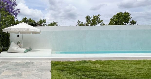 A minimalist outdoor pool area with a long turquoise swimming pool enclosed by a translucent pale glass privacy wall. A white umbrella shades a low white daybed with pale cushions beside the pool. The pool deck uses irregular light grey stone paving, bordered by a neatly maintained lawn and mature greenery under an overcast sky.