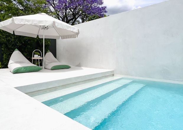 A bright minimalist outdoor pool courtyard with a turquoise rectangular swimming pool, broad white rendered walls and paving, and shallow entry steps. A white cantilever-style umbrella shades two white-and-green loungers beside a small side table, while leafy trees and purple flowering branches rise above the courtyard wall.