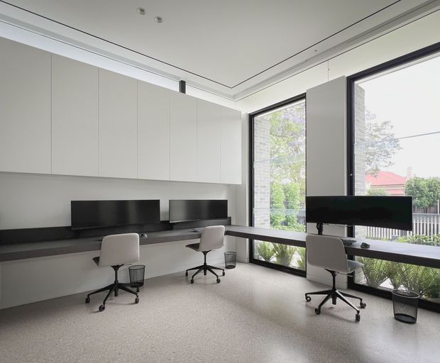Bright, minimalist home office with a long built-in desk running along the wall and beneath large corner windows. Three compact workstations feature black monitors, white swivel chairs, and small wastebaskets. The room has pale built-in overhead cabinetry, dark desk surfaces, a light speckled floor, slim dark window frames, and concealed linear ceiling detailing. Trees and neighbouring buildings are visible outside.