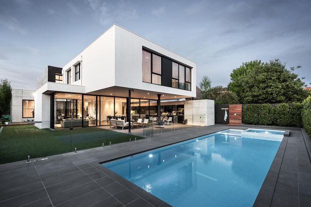 Exterior architectural photograph of a large two-storey contemporary home with crisp white rendered walls, extensive dark-framed glazing, a covered outdoor entertaining area, dark paved pool terrace, and a rectangular swimming pool. The landscaped setting includes hedges and lawn, with a moody overcast sky.