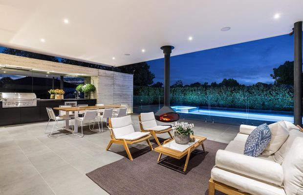 A sophisticated covered outdoor entertaining area at dusk, featuring an outdoor kitchen and dining zone, a central suspended fireplace, lounge seating, large-format tiled flooring, and a glass balustrade overlooking landscaped greenery and a lit pool or water feature.