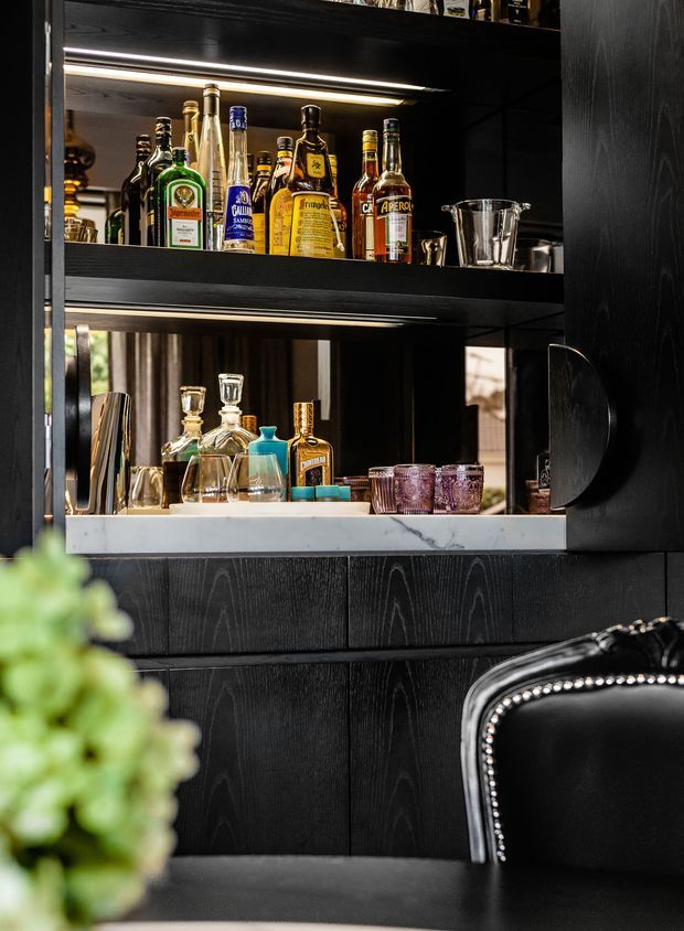 A close-up of a sophisticated built-in home bar with black vertical-grain cabinetry and open illuminated shelving. The shelves display liquor bottles, decanters, barware and coloured tumblers, with a mirrored backing and a light marble or stone ledge. A black leather chair with metallic nailhead trim is partially visible in the foreground.