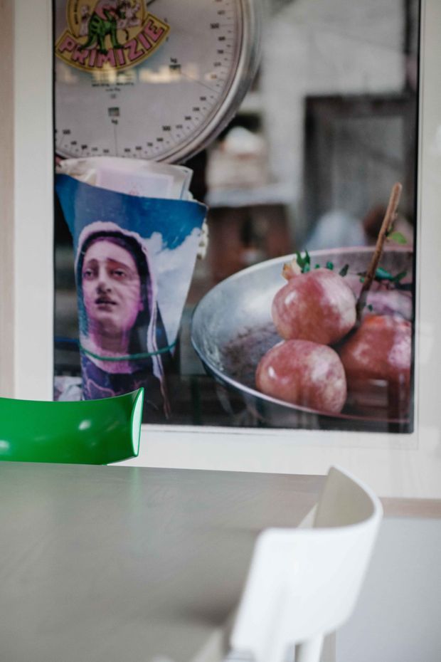 A close-up view of a contemporary dining area with a grey tabletop, white molded dining chairs, and a bright green chair partially visible. A large framed artwork or photograph depicting a bowl of apples and a kitchen scale hangs on the wall, adding a colourful eclectic accent.