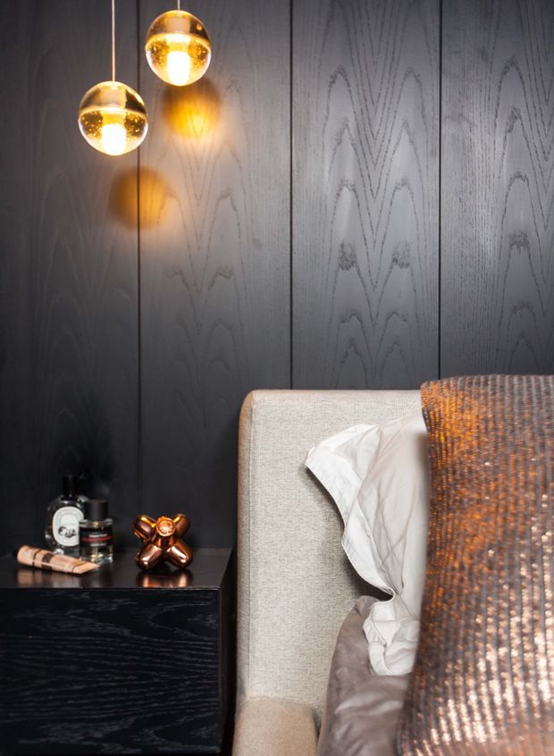 Close-up of a sophisticated bedroom corner featuring a tall upholstered bedhead in pale textured fabric, dark vertically panelled timber wall cladding, a matching dark bedside cabinet, and two warm glowing spherical pendant lights. Neutral bedding and a textured brown-grey cushion create a refined, moody atmosphere, with small decorative objects on the bedside table.