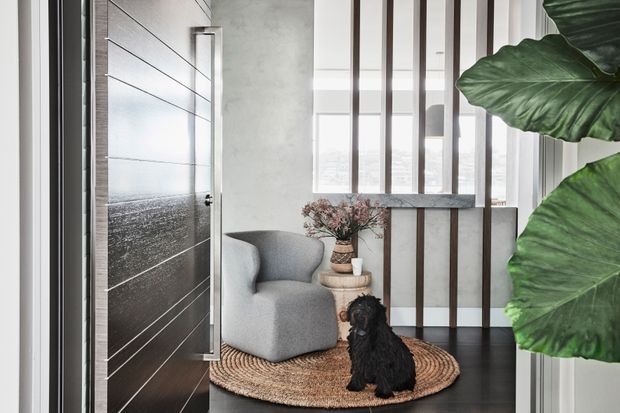 A refined residential entryway with a large dark timber pivot door, a grey upholstered lounge chair, a woven round rug, a small stacked-stone side table with vase, and a black dog. Vertical timber slats frame a bright window, while large tropical leaves partially screen the view on the right. The space has a contemporary, calm, natural-material palette.