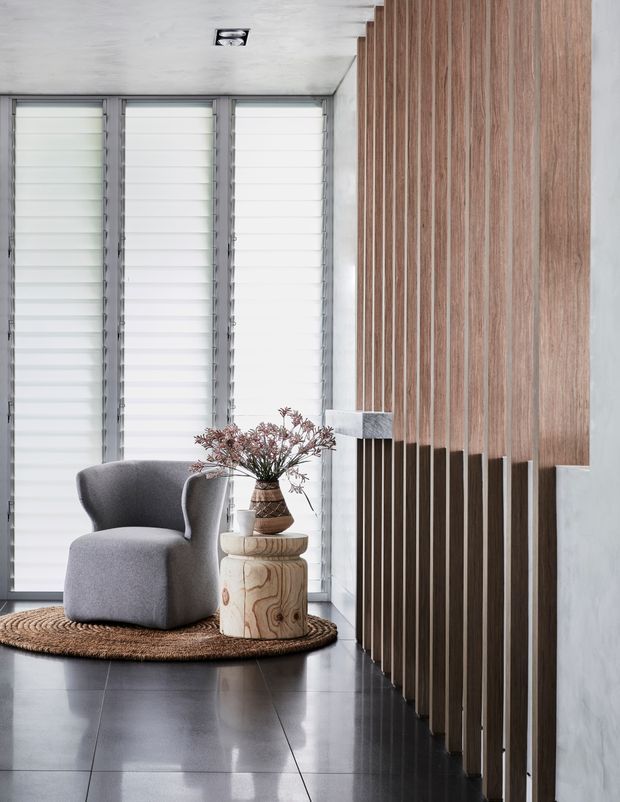 A refined modern interior vignette featuring a sculptural grey armchair beside a small round timber side table, set on a woven natural-fibre rug. Floor-to-ceiling vertical timber battens form a strong architectural feature wall and partial screen, contrasted with pale concrete walls, translucent louvred glazing, and dark polished flooring. A small branch arrangement adds an organic accent.