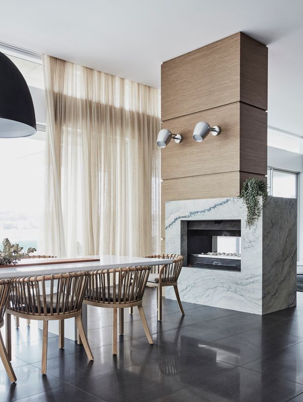 A bright contemporary dining area with a long dining table and woven timber-framed chairs beside a dramatic marble-clad fireplace. Floor-to-ceiling sheer curtains cover large windows, while a tall wood-panelled feature wall rises to the ceiling above the fireplace. Dark polished floor tiles, sculptural pendant lighting, and restrained neutral furnishings create a refined, airy interior.