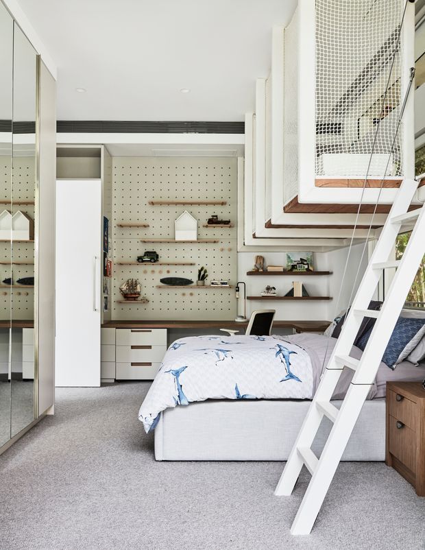 Bright, compact bedroom with a raised loft bed accessed by a white ladder. The room features built-in cabinetry, a desk, open shelving, pegboard storage, mirrored wardrobe doors, and a small collection of decorative objects. Neutral white and light-grey finishes are complemented by timber shelving and blue patterned bedding.