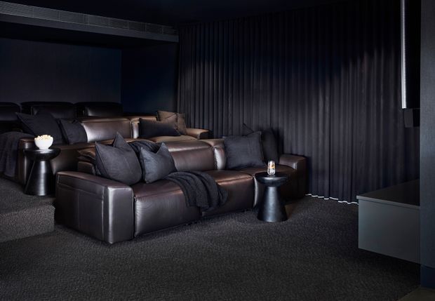A dark, sophisticated home cinema/media room with tiered rows of deep brown leather reclining sofas, black and charcoal cushions, small sculptural side tables, dark carpet, and full-height black drapery. A large screen is partially visible at right, while subtle linear ceiling detailing and concealed lighting reinforce the cinematic atmosphere.