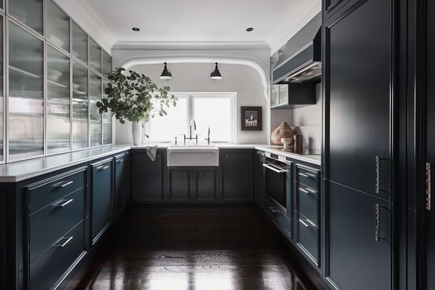 A refined galley-style kitchen with dark navy or charcoal cabinetry, polished dark timber flooring, a white farmhouse sink, stainless steel appliances, and a large industrial-style glazed partition on the left. The room features a bright window, decorative wall moulding with curved detailing, pendant lights, and a restrained monochromatic palette.