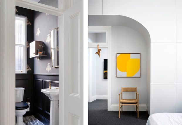 A refined interior view looking from a bright bedroom or sitting area into a compact powder room. The powder room features dark painted wall panelling, patterned wallpaper above the dado, a white pedestal basin, toilet, wall mirror and narrow window. The adjacent room has white paneled walls, a dramatic curved arched opening, grey carpet, a timber-framed chair, contemporary yellow artwork and the edge of a bed.
