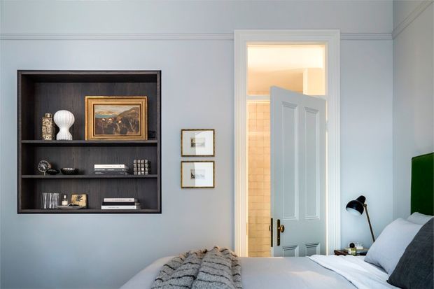 A serene bedroom with pale blue walls, decorative wall moulding, a dark built-in display shelf, framed artwork, and a partially open paneled door leading to a tiled bathroom. The bed occupies the foreground with layered grey and white bedding, while a black bedside lamp and green upholstered headboard add contrast.
