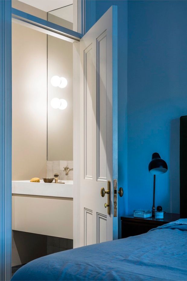 View from a blue-toned bedroom through an open panelled door into a compact bathroom. The bathroom has a floating white vanity with integrated basin, wall mirror, round wall lights and dark tiled flooring. The bedroom includes a blue bedspread, vivid blue walls, a dark bedside table and black lamp. White traditional-style door trim contrasts with the saturated blue interior.