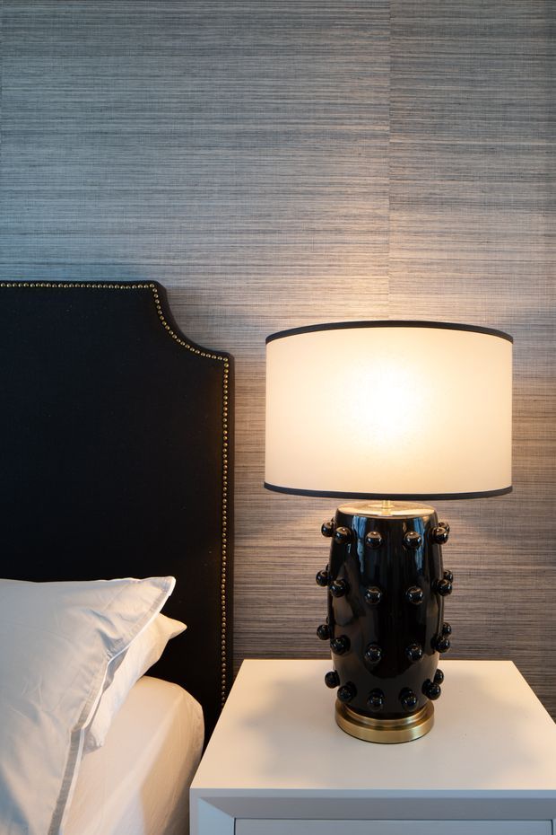 Close-up view of a refined bedroom corner featuring a black upholstered bedhead with brass nailhead trim, white bedding, a white bedside table, and a distinctive glossy black table lamp with a warm cream shade. The wall is finished in horizontally textured grey-brown wallpaper or paneling, creating a sophisticated, moody atmosphere.