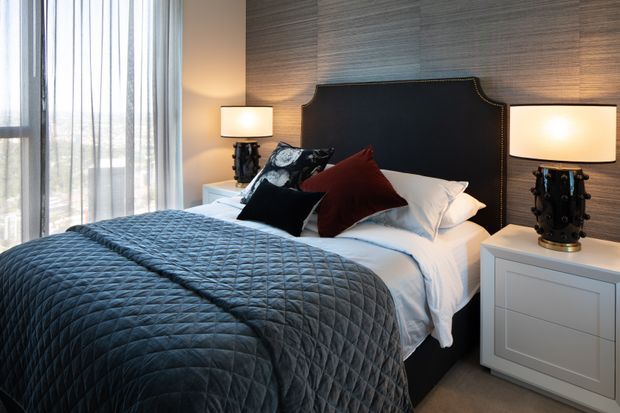 A sophisticated bedroom with a large upholstered bed, dark quilted coverlet, layered white and burgundy pillows, and matching bedside lamps. The bed is set against a textured grey feature wall with a dark, nailhead-trimmed headboard. Floor-to-ceiling sheer curtains provide soft natural light, while white bedside cabinetry and neutral carpeting complete the space.