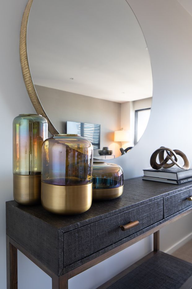 A styled entryway or hallway console vignette featuring a dark textured console table with timber legs and drawer pulls, an oversized round mirror with a woven natural frame, iridescent glass vessels with metallic bases, and a sculptural looping ornament. A living area is visible in the mirror reflection.
