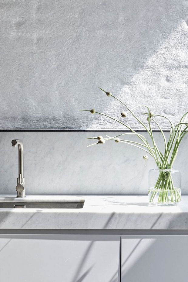 Bright close-up of a minimalist kitchen sink area with a slim metal tap, pale marble-look benchtop and splashback, smooth light cabinetry, and a clear glass vase of delicate greenery. Strong sunlight creates diagonal shadows across the white surfaces.
