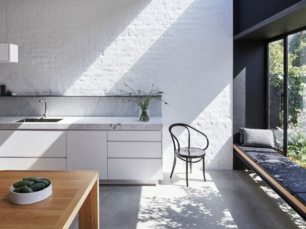 A bright, minimalist kitchen with white textured masonry walls, sleek pale cabinetry, a stone benchtop, and a large black-framed window. Strong sunlight creates diagonal shadows across the wall and polished concrete floor. A timber dining table, simple black chair, and built-in window seat add warmth and contrast.