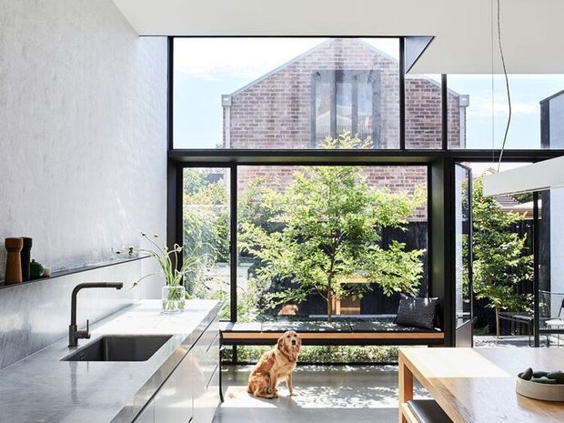 Bright contemporary kitchen with a long pale stone island and integrated sink, dark tapware, minimalist cabinetry, and extensive black-framed floor-to-ceiling glazing. The glazing overlooks a leafy courtyard and reveals a brick gabled building beyond. A dining table and seating are partially visible at right, with a dog in the courtyard.