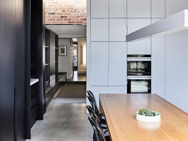 A contemporary open-plan kitchen and dining area with a long light timber dining table, black sculptural chairs, dark built-in cabinetry, pale grey full-height kitchen joinery, integrated appliances, and a large angular rangehood. Polished concrete flooring, exposed brickwork, and a dark metal-framed transition toward a hallway create an industrial character.