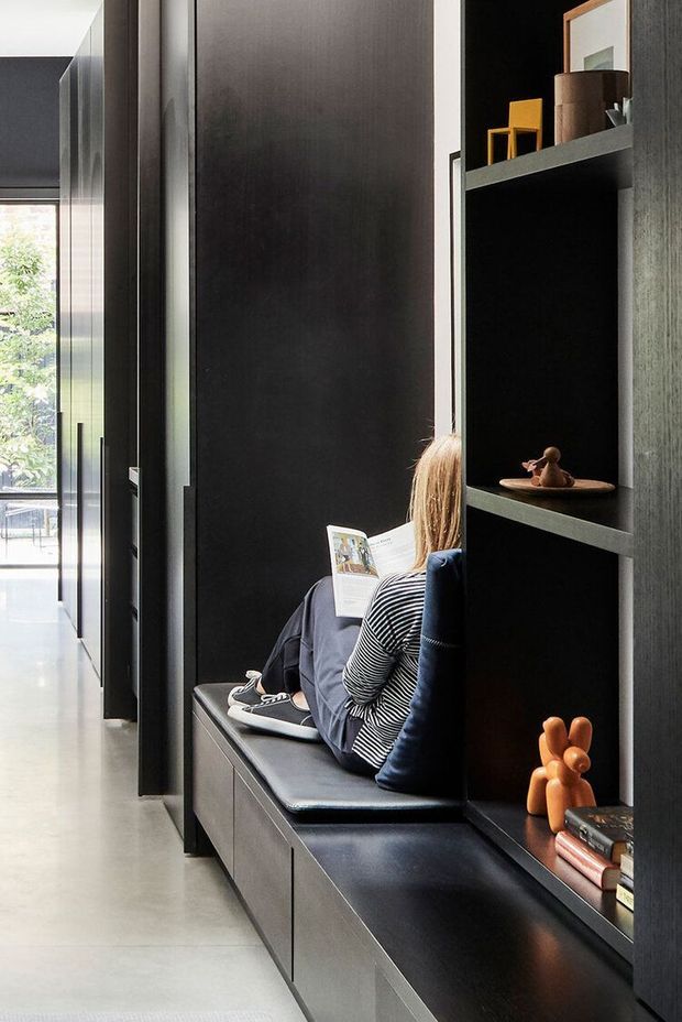 A narrow, sophisticated interior corridor featuring dark full-height built-in cabinetry and open display shelving. A long integrated bench with dark upholstery runs along the joinery, creating a compact reading or seating nook occupied by a person. The space has a polished concrete or light grey floor, strong black and charcoal tones, and glimpses of a bright garden-facing area through glazing at the end.