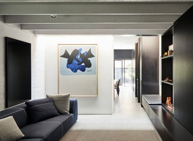 A contemporary living room with a dark sectional sofa, wall-mounted television, built-in dark timber entertainment shelving, and a framed abstract artwork. The room has a bright neutral palette, exposed painted ceiling beams, and a view through the hallway toward a dining area and glazed exterior opening.