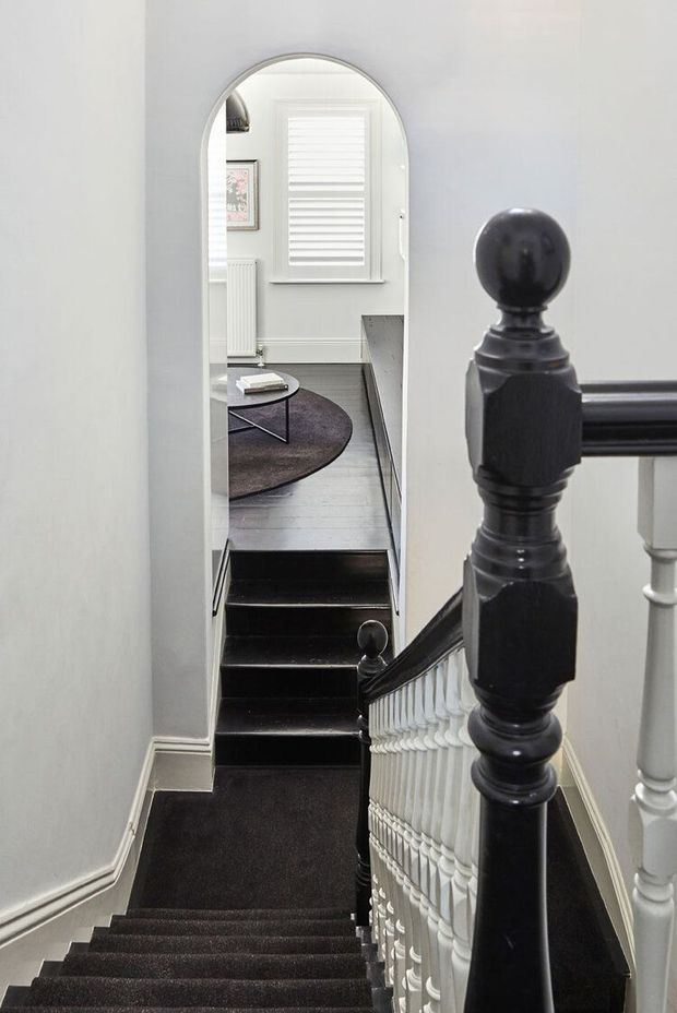 A view looking down a traditional residential staircase with dark carpeted steps, white decorative balustrades, and a prominent dark timber newel post. An arched opening leads to a lower-level living area with a round rug, small coffee table, framed artwork, radiator, and plantation-shuttered window. The space has a refined monochrome palette with classic detailing.