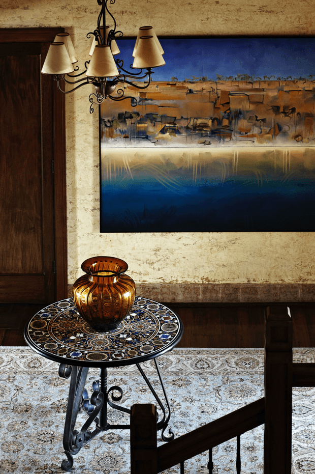 A richly styled traditional interior vignette featuring dark timber wall panelling and door, textured cream plaster, a large blue and ochre abstract artwork, an ornate wrought-iron chandelier, and a small round decorative mosaic-topped table with an amber glass vase. A patterned pale rug and timber stair railing are visible in the foreground.
