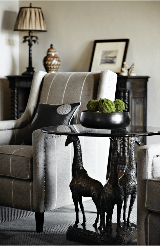 A refined traditional sitting area featuring two upholstered armchairs, a glass-topped side table with sculptural giraffe-leg supports, and a black bowl of moss-like spheres. A dark wood console with a decorative lamp, vase, framed artwork, and ornaments sits against a pale neutral wall. The room has tailored upholstery, nailhead trim, patterned carpet, and an elegant, layered decorative scheme.