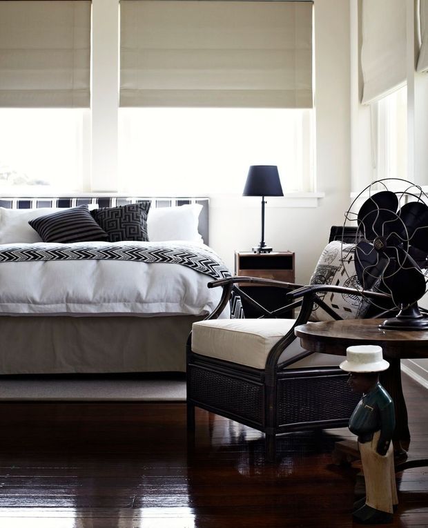 A refined bedroom with a white upholstered bed dressed in graphic black-and-white bedding, dark polished timber flooring, and large windows fitted with beige Roman blinds. A black table lamp, woven dark armchair, round side table, sculptural fan, and decorative figurine add layered character to the room.
