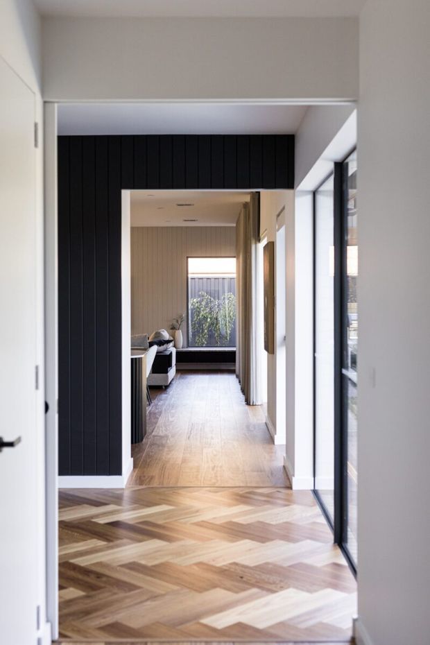 A bright, narrow residential hallway or entry passage viewed through open white doors. The foreground has a distinctive natural timber herringbone parquet floor, leading toward a light-filled living area with seating and a large window. A dramatic black vertically panelled feature wall frames the opening, while the right side has full-height dark-framed glazing and white walls. The interior has a refined contemporary feel with strong contrast between the black wall, white surfaces, and warm timber flooring.