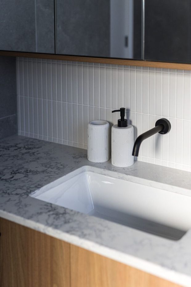 Close-up of a refined bathroom vanity featuring a white rectangular undermount basin, grey veined stone benchtop, warm timber cabinetry, slim white vertically stacked tile splashback, dark charcoal overhead cabinetry, and matte black wall-mounted tapware. White textured soap and tumbler accessories sit beside the basin.