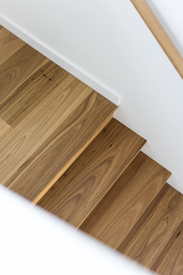 Close-up view of a straight staircase finished in warm natural timber, with prominent wood grain on the treads and white painted walls and trim surrounding the stairwell. A simple timber handrail is partially visible at the upper right.