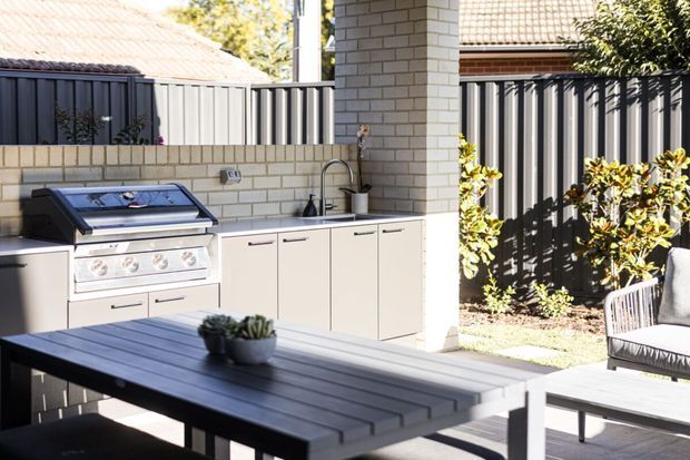 Bright outdoor entertaining area with a built-in barbecue kitchen, stainless-steel grill, sink and light cabinetry set against a pale brick wall. A dark slatted dining table, chairs, and planted boundary garden complete the contemporary courtyard setting.